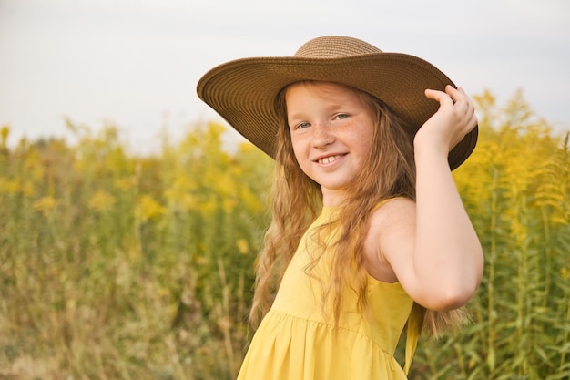 노란 드레스와 모자를 쓴 행복하고 웃는 귀여운 소녀가 들판을 걷습니다. 여름 야외 생활 방식