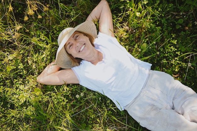 Happy smiling elderly senior woman in straw hat having fun lying on grass posing in summer garden