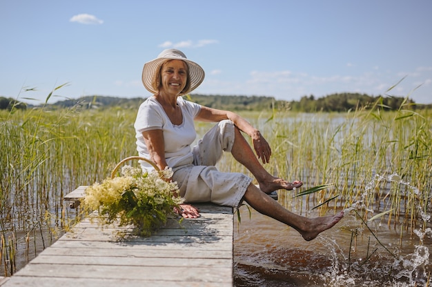 Happy smiling elderly senior woman in straw hat sitting on wooden pier by lake with flowers in