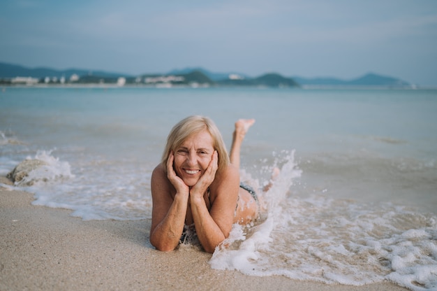 Happy smiling excited elderly senior woman tourist playing in water and swimming in the big waves on the ocean sea beach. Traveling along Asia, active lifestyle concept.