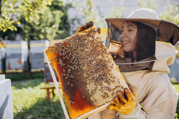 Happy smiling female Beekeeper in protective suit holding honeybee frame with bees at apiary