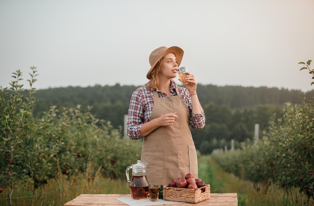 가을 수확 시간 동안 과수원 정원에 서 있는 유리에 맛있는 사과 주스를 마시는 행복한 미소 여성 농부