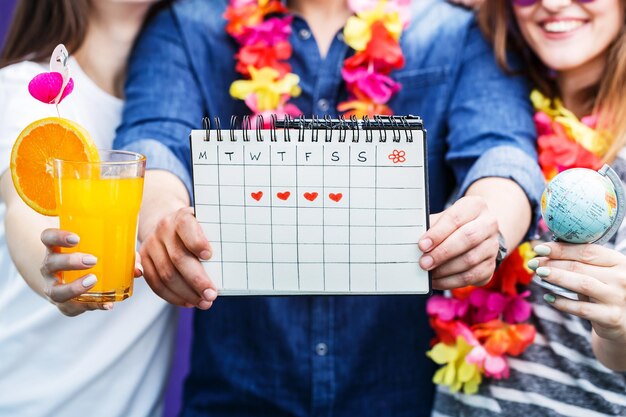 Photo happy smiling friends two girls and bearded man holds vacation trip calendar globe and orange drink