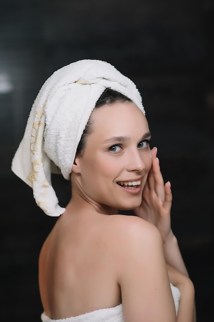 Foto felice ragazza sorridente applicare crema viso guardando la fotocamera bella signora sana avvolta in asciugamani mettere idratante crema giorno nutriente lifting sulla morbida pelle idratata