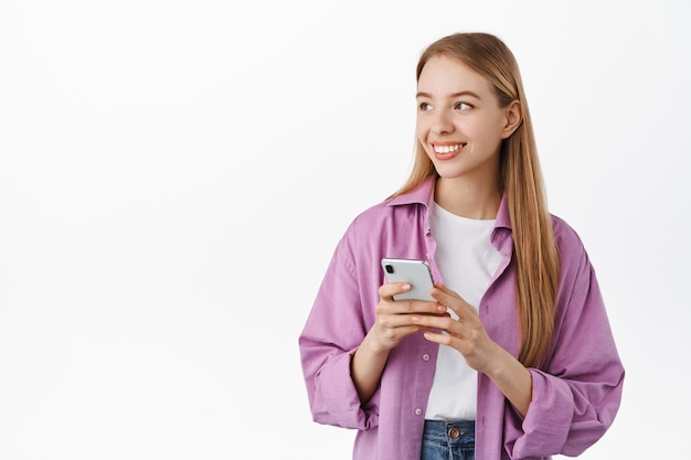 Ragazza sorridente felice in abiti casual, usa lo smartphone, tiene il telefono in mano e guarda da parte il logo con il viso allegro e rilassato, in piedi sul muro bianco