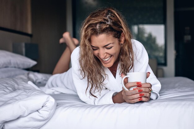 Счастливая улыбающаяся девушка в белом теплом халате, лежа на кровати с чашкой в руках. Уютное зимнее настроение.