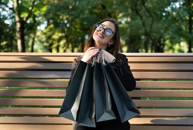 Happy smiling girl with black shopping bags on bench in park Successful shopping