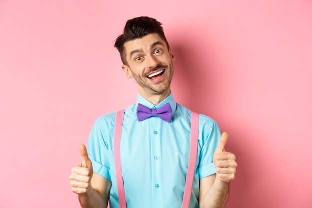 Happy smiling guy in bow-tie and suspenders showing thumbs up, praising good job, approve nice work, standing over pink satisfied.