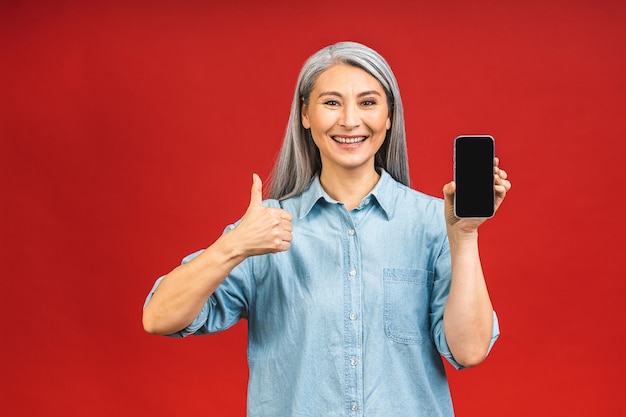 Felice sorridente felice donna matura senior in casual che mostra lo schermo vuoto dello smartphone mentre si guarda la fotocamera isolata su sfondo rosso utilizzando il telefono