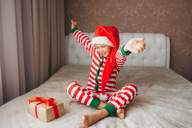 Happy smiling joyful child boy in Santa Claus hat, holding a Christmas present in his hands, sitting on the bed.