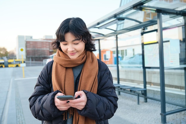 スマートフォンを見てバス停に立っている携帯電話を使用して幸せな笑顔の韓国の女の子