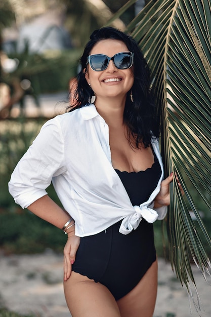 Happy smiling lady in a black bodysuit and white sweater wearing sunglasses posing next to palm leaves. Against the background of the jungle. Summer concept