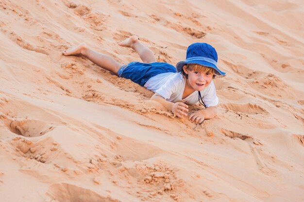 happy smiling little boy in hat rolls down the sand dune in the desert