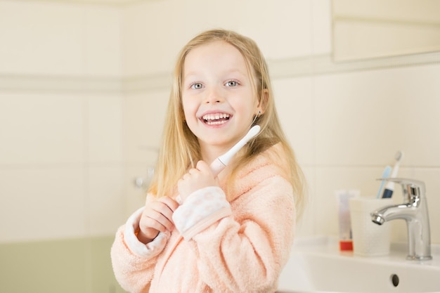 Bambina sorridente felice lavarsi i denti usando uno spazzolino elettrico ad ultrasuoni in bagno a casa orale ...