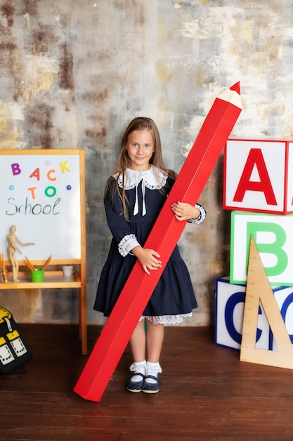 Happy smiling  little girl in dress stands with large pencil in her hands at school. Back to school