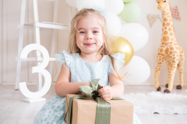 Happy smiling little girl holds gift box on balloons background birthday party