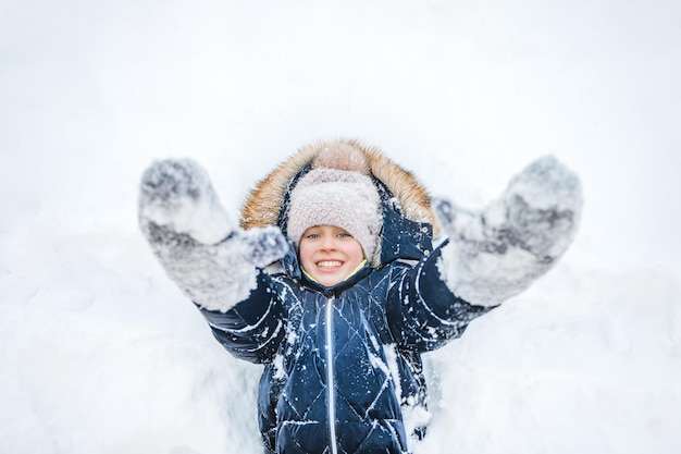 A happy smiling little girl lies in the snow with her arms outstretched to the camera in winter