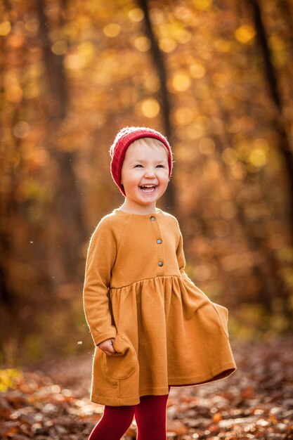 A happy smiling little girl in the red knitted hat walking in the autumn forest