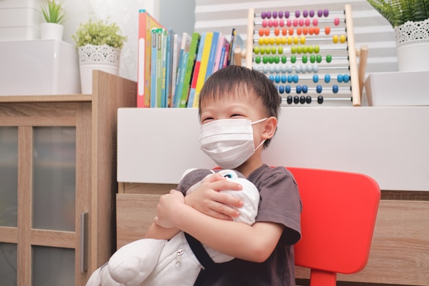 Happy smiling little kindergarten Asian boy child hugging his dog plush toy  both in protective medical masks