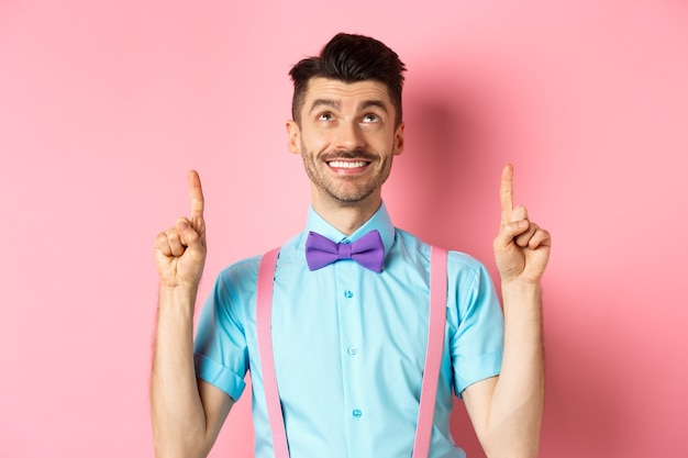 Happy smiling man in bow-tie pointing, looking up, showing something cool, standing on pink.