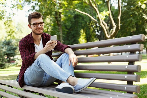 Happy smiling man in glasses with smartphone outdoors. Handsome casual boy checking email and surfing web on mobile, sitting in park with legs raised on bench, copy space