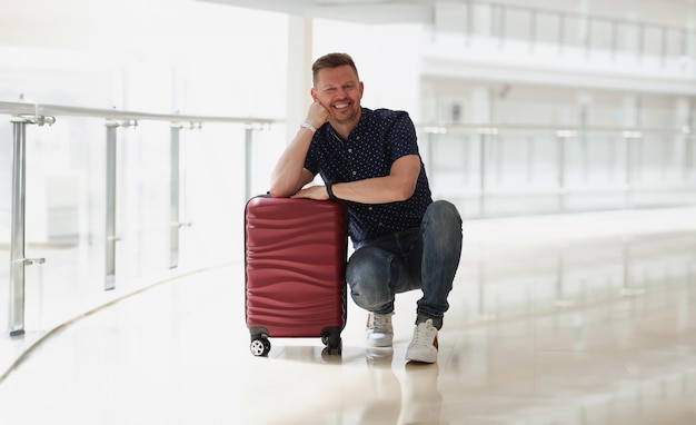 Счастливый улыбающийся мужчина, позирующий возле чемодана, планирующий поездку за границу, ждет отъезда в аэропорту