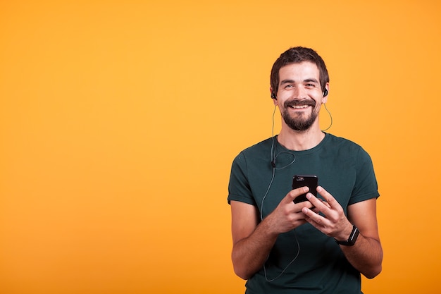 Uomo sorridente felice con le cuffie sull'ascolto di musica sul suo smartphone isolato su sfondo giallo. stile di vita dell'intrattenimento mobile