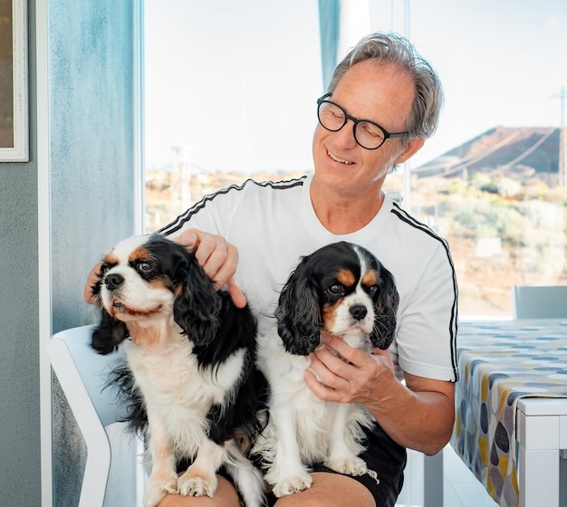 Happy smiling mature man cuddling his two cavalier king Charles spaniel dogs sitting on his legs