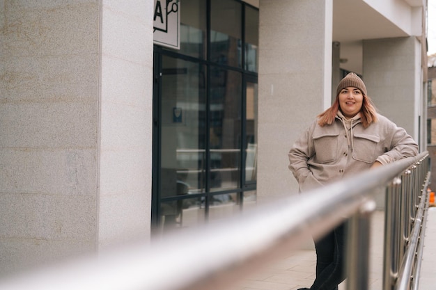 Donna in sovrappeso sorridente felice con cappello caldo e giacca in piedi in posa vicino alla ringhiera dell'edificio per uffici in una strada cittadina in una nuvolosa giornata autunnale