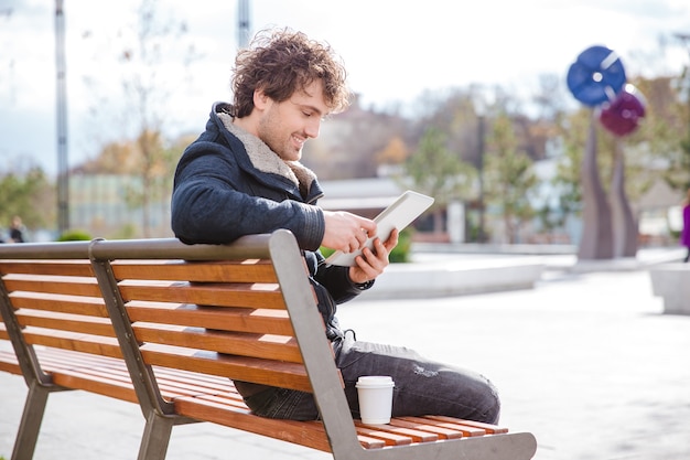 Sorridenti contenti contenti giovane maschio riccio seduto su una panchina nel parco usando tablet e bevendo caffè