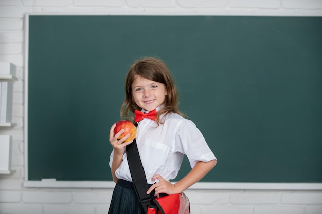 Happy smiling preteen girl kid schoolgirl with wears schhol uniform holding backpack on blackboard background looking at camera Education learning and children concept