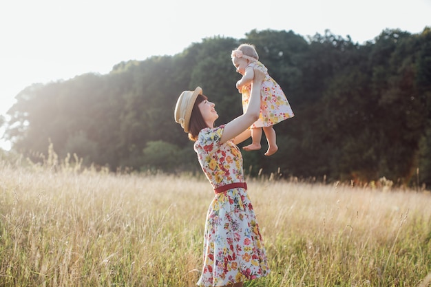 Happy smiling pretty mother wearing dress and straw hat, holding and raising up little baby daughter playing in beautiful summer field