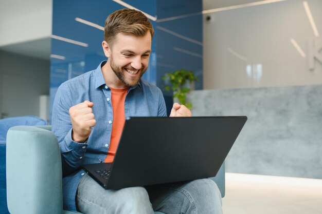 Happy smiling remote online working man in casual outfit with laptop in joyful successful winning gesture sitting in an coworking office at a work desk