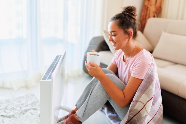 Happy smiling satisfied joyful young woman with plaid drinking hot tea and warming near portable electric heater at home