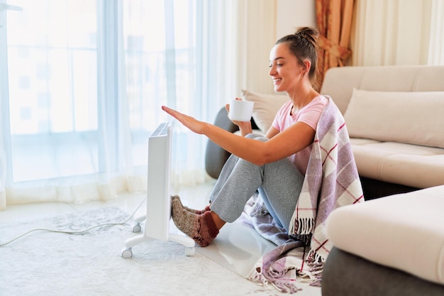 Happy smiling satisfied joyful young woman with plaid and\
knitted socks drinking hot tea and warming near portable electric\
heater at home