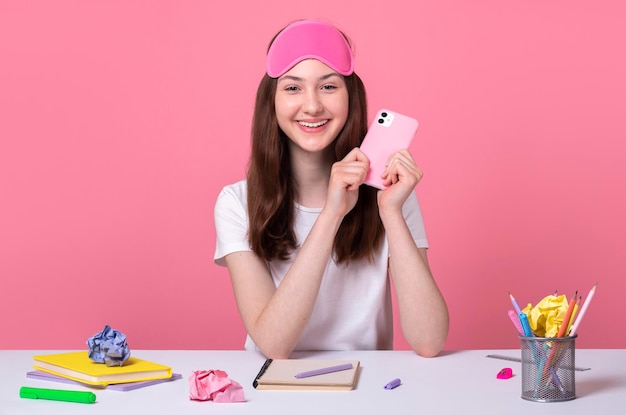 Happy smiling school girl sits table desk and holding mobile smart phone in hands on pink background