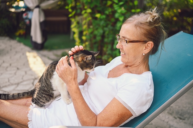 国内のぶち猫を抱き締める屋外の夏の庭でリラックスしたメガネで幸せな笑顔の年配の女性。引退した老人と動物のペットの概念