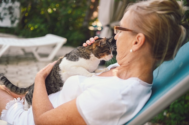 国内のぶち猫を抱き締める屋外の夏の庭でリラックスしたメガネで幸せな笑顔の年配の女性。引退した老人と動物のペットの概念