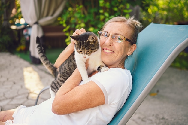 Happy smiling senior elderly woman in glasses relaxing in summer garden outdoors, hugging domestic tabby cat