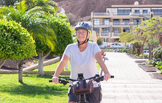 Happy smiling senior woman riding with her electro bike in public park enjoying healthy lifestyle Elderly woman with eyeglasses wearing helmet