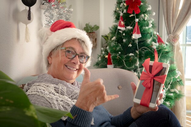 Happy and smiling senior woman wearing a Santa Claus hat holding a digital tablet as Christmas present - merry Christmas at home for an elderly retired people enjoying holidays