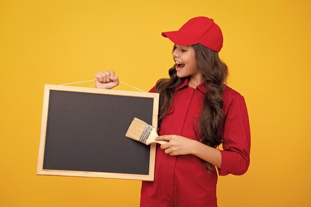Happy smiling teenager girl builder in building uniform and cap Painter with painting brush tool or paint roller Worker hold blackboard isolated on yellow background Kids renovation concept