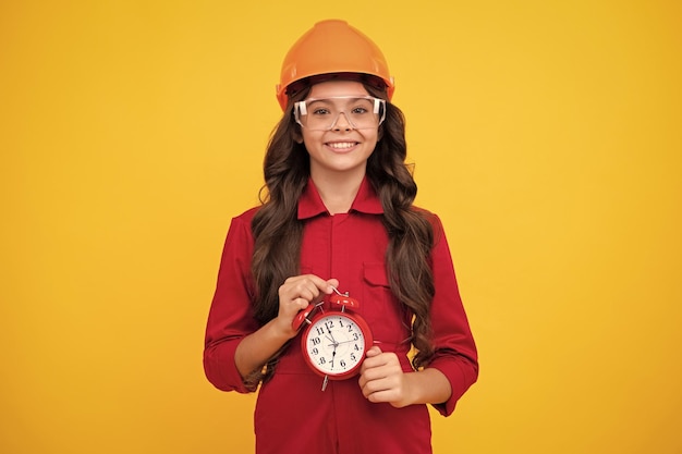 Happy smiling teenager girl in builder helmet and clock alarm time to renovation and repair child in hard hat kid builder