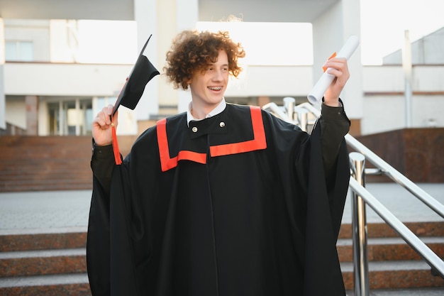 Happy smiling university graduate in mantle holding diploma in\
raised hand and expressing happiness over university building at\
background successful graduating from university or college