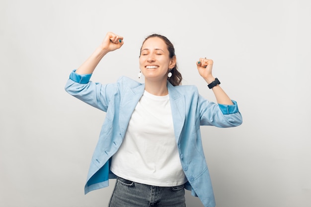 Happy smiling winner lady is dancing over white background with hands up.