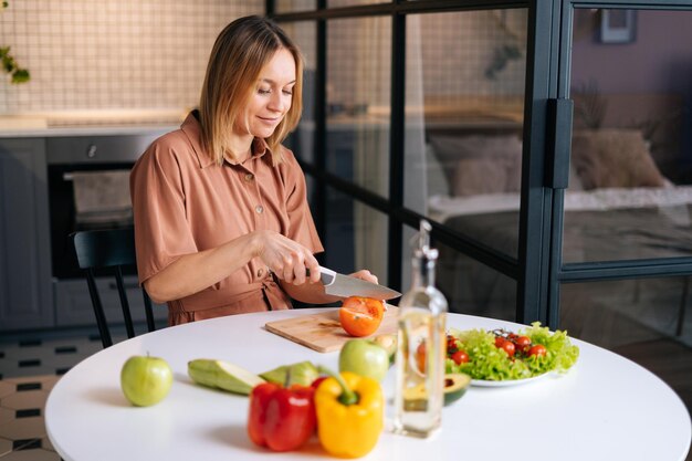 モダンなキッチンで野菜サラダ用のナイフで新鮮な有機トマトを切る幸せな笑顔の女性。笑顔のスリムな菜食主義の女性は、健康的なビーガンディナーの食事を準備します。健康的な食事の概念。