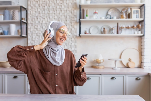 Happy and smiling woman dancing and singing at home in kitchen muslim woman in hijab and headphones