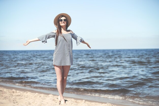 Happy smiling woman in free bliss on ocean beach standing with open hands. Brunette female model in sunglasses and hat enjoying nature during travel holidays vacation outdoors, medium shot.