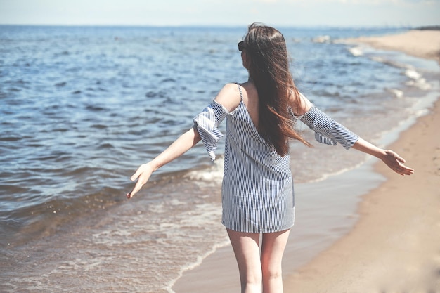 두 손을 벌리고 바다 해변에 서서 자유로운 행복을 누리며 웃고 있는 행복한 여성. 야외에서 여행 휴가를 보내는 동안 자연을 즐기는 여름 드레스를 입은 갈색 머리 여성 모델의 초상화.