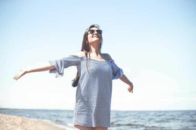 Happy smiling woman in free bliss on ocean beach standing with open hands. Portrait of a brunette female model in summer dress enjoying nature during travel holidays vacation outdoors.